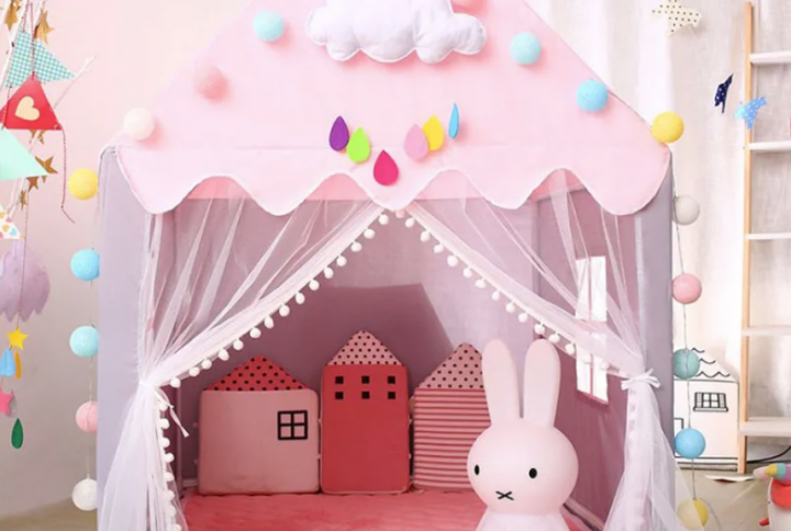 Tipi rose décoré avec des pompons colorés, des nuages et des gouttes de pluie en feutrine, avec des coussins en forme de maisons et une lampe en forme de lapin à l'intérieur, créant un espace de jeu magique pour enfants.
