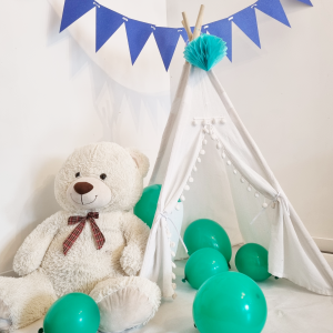 Tipi pour enfant en tissu blanc avec pompons, décoré d’un ornement bleu sur le sommet, accompagné d’un ours en peluche et de ballons verts, avec une guirlande de fanions bleus en arrière-plan.