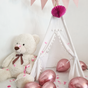 Tipi pour enfant en toile blanche avec pompons et guirlande de fanions roses, décoré de ballons roses et de confettis en forme de cœur. Un ours en peluche géant est assis à l'entrée.