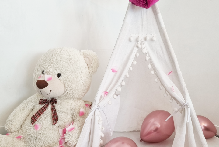 Tipi pour enfant en toile blanche avec pompons et guirlande de fanions roses, décoré de ballons roses et de confettis en forme de cœur. Un ours en peluche géant est assis à l'entrée.
