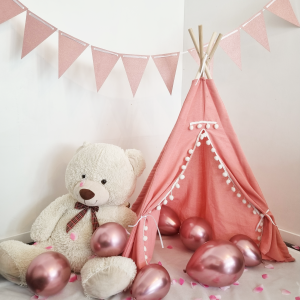 Tipi pour enfant en tissu rose avec pompons blancs, accompagné d’un ours en peluche, de ballons roses métallisés et de pétales, avec une guirlande de fanions roses en arrière-plan.
