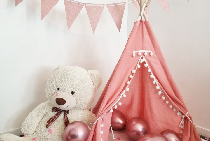 Tipi pour enfant en tissu rose avec pompons blancs, accompagné d’un ours en peluche, de ballons roses métallisés et de pétales, avec une guirlande de fanions roses en arrière-plan.